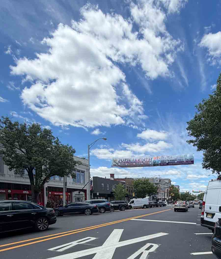 A street view of downtown Englewood, NJ, featuring a Juneteenth Independence Day banner hanging across the road under a cloudy blue sky_-o2